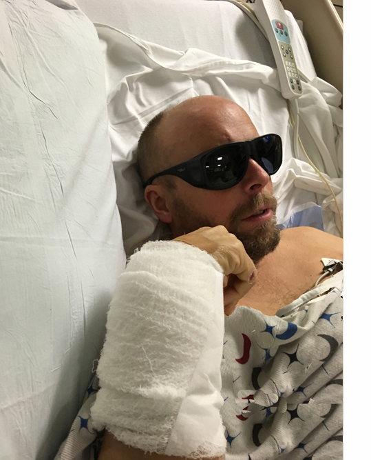 Paul is lying propped up in the hospital bed, wearing dark wraparound glasses.